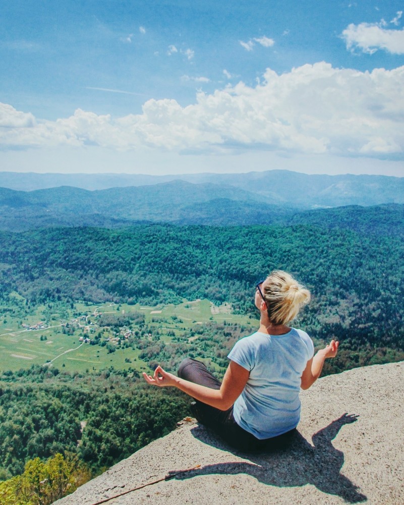 A woman in a zen state of mind sitting on a cliff with mountains in the background